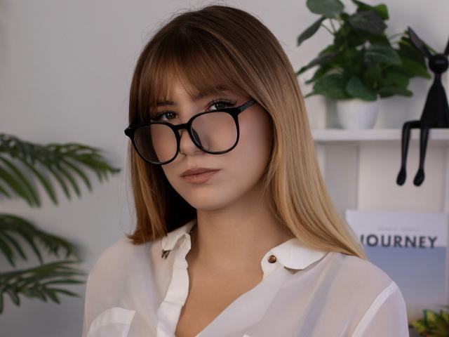 ChloeKingsley with straight blonde hair, black-rimmed glasses, and a white button-up shirt, showing off her cute face and small, perky tits. Background includes green plants and white shelves.