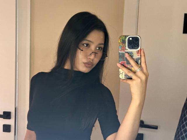 LeonilaSeewald with long black hair, black turtleneck, and face paint marks, taking a mirror selfie with a colorful phone case, showing off ringed fingers.