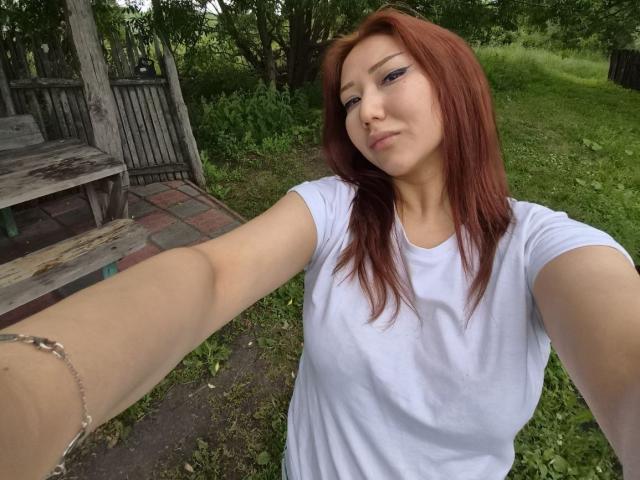 SheritaAshkettle with red hair, white t-shirt, and eyeliner, taking a selfie outdoors. She's extending her arm, wearing a bracelet, with a slight smirk. Her tits are visible.