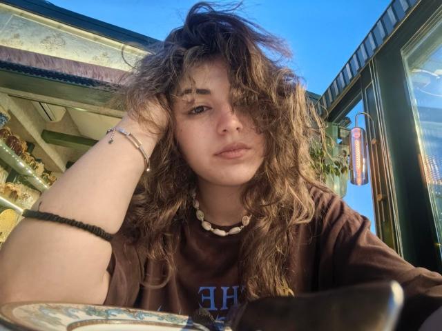 JeneeDetherage with messy brown curly hair, brown t-shirt, white bead necklace, black bracelet, resting chin on hand, looking tired, sitting outside, blue sky, sunny.