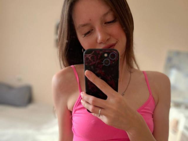DollieSkweres with brown hair, pink top, holding phone, taking selfie, smiling, small zit on cheek, pink nipples, ring on finger, phone case with dark red pattern.