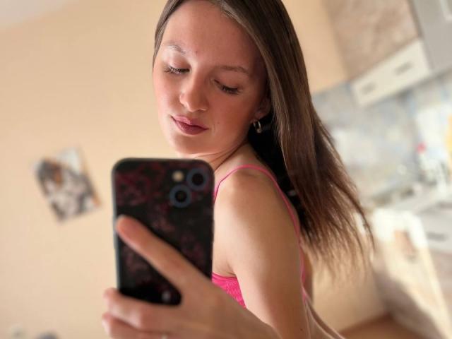 DollieSkweres with brown hair, pink tank top, taking a selfie with a black phone in a blurry room, showing her ass and smiling.
