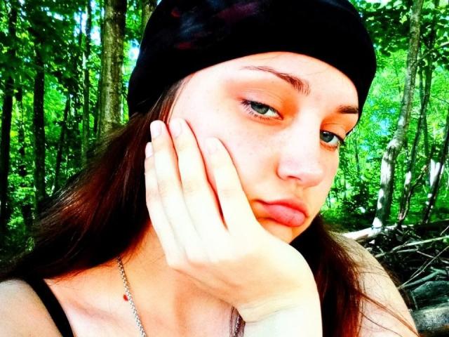 Georgina Brockwell mit einer schwarzen Mütze, Hand auf ihrer leicht hochgezogenen Wange, grüner Wald im Hintergrund, helle Haut, braunes Haar, hellrosa Lidschatten, dezenter Eyeliner, Halskette mit roter Perle..