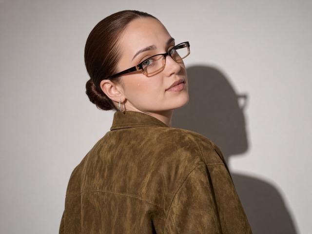 Georgina Brockwell mit braunen Haaren zu einem Dutt gebunden, trägt eine Brille, eine grüne Wildlederjacke und goldene Ohrringe, sie schaut über ihre Schulter, ihr Schatten ist an der Wand zu sehen..
