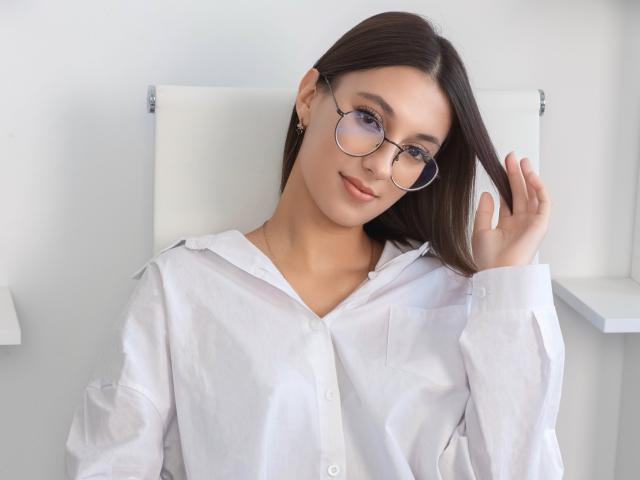 Emma Everett, con gli occhiali, una camicia bianca, capelli castani e un sorriso carino, che si tocca i capelli, mostrando leggermente il seno attraverso la camicia, in una stanza luminosa e pulita..