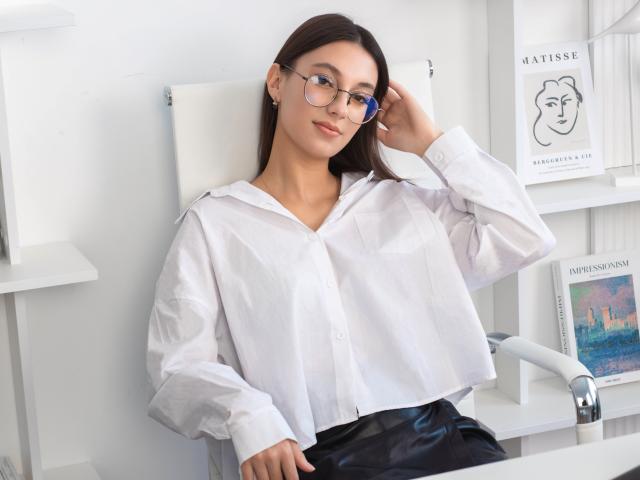 Emma Everett mit Brille, weißem Hemd und schwarzem Lederrock, sitzt auf einem Stuhl, die Hand im Haar, wirkt entspannt, niedlich und attraktiv, mit dezentem Make-up und langen braunen Haaren..