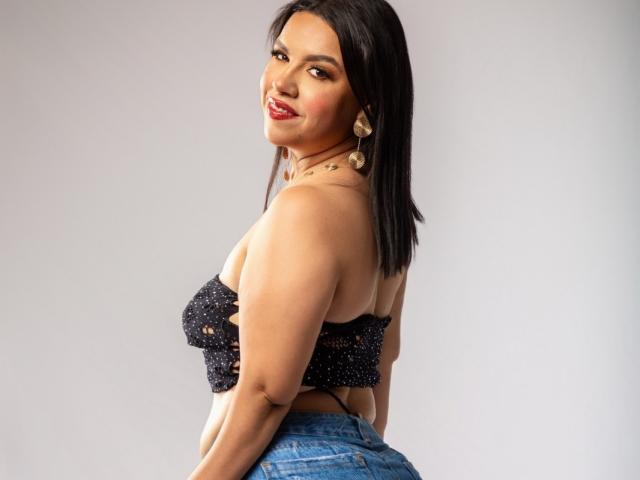 AlannaSloppy with dark hair, smiling, wearing a sparkly black strapless top, and blue jeans, showing her curvy ass and medium-sized tits, with gold earrings.