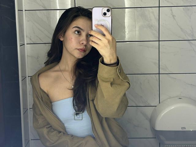 Anne Kofron, met lang bruin haar, draagt een beige jas over een blauwe top zonder bandjes, en maakt een spiegelzelfportret met een witte telefoon, waarbij haar gezicht en bovenlichaam te zien zijn..