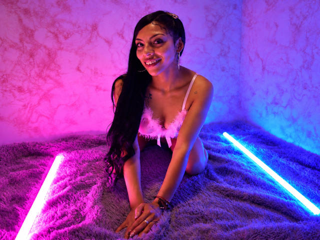 Roxanna Moretti, avec ses longs cheveux noirs, porte un soutien-gorge en plumes roses, assise sur une couverture violette et duveteuse, éclairée par des néons roses et bleus, et sourit à l'appareil photo..