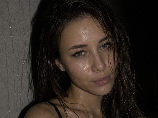 AmmaLoyXX has wet brown hair, glistening skin, and blue eyes, looking slightly annoyed. She's wearing a black top, and her tits are partially visible. Dark background.