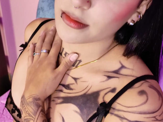 SophiieBlair with black hair, red lipstick, nose ring, and tattoos, wearing a black lace bra, touching her chest, showing her tits and tattoos, with a gold necklace.