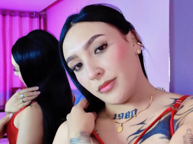 SophiieBlair with black hair, nose ring, and tattoos, wearing red lingerie, looking at camera, mirror reflection showing back, pink lighting, gold necklace, and tattoo on chest.