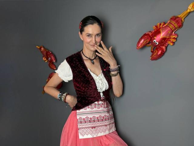 TayaFlavy, mit dunklem Haar, trägt eine rote Samwesten, eine weiße Bluse, einen pinken Rock und mehrere Armbänder. Sie steht mit einem Finger am Mund, umgeben von roten Krabben-Ballons..