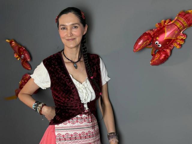 TayaFlavy mit geflochtenen Haaren, die eine rote Sammweste, eine weiße Bluse, einen rosa bestickten Rock und Armbänder trägt, steht vor einer grauen Wand mit roten Krabben-Ballons..