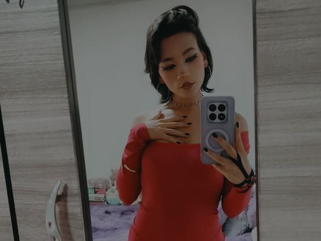 HenutmireRossi with short black hair, wearing a red off-shoulder dress, holding a phone, hand on chest, black nail polish, mirror selfie, bedroom, choker necklace.