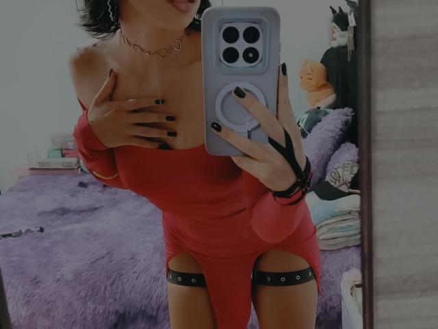 HenutmireRossi taking a mirror selfie, wearing a red dress with garter straps, black nail polish, and a chain necklace, covering her tits with one hand, showing a big cock.
