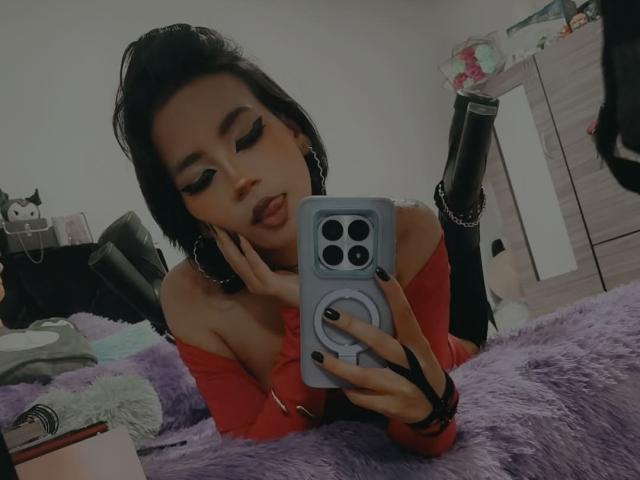 HenutmireRossi with dark hair, red off-shoulder top, black nails, and hoop earrings, taking a mirror selfie with a blue phone, resting on a purple furry surface.
