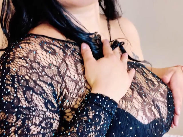 AntonelaWayne in black lace top with her hand on her tits, showing her large, bare breasts through the see-through pattern. Her black hair is down, and her skin is tan.