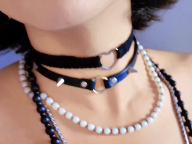 JadeMilleer's neck, wearing two black leather chokers with heart and ring metal accents, and multiple bead necklaces—white, black, and blue—on a light-skinned, slightly tanned, curly-haired person.