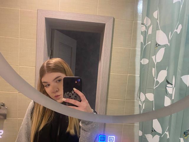 NorahHabrock with blonde hair, taking a mirror selfie in a bathroom, holding a black phone, wearing a gray shirt, looking slightly pouty, with a green leaf-patterned curtain.