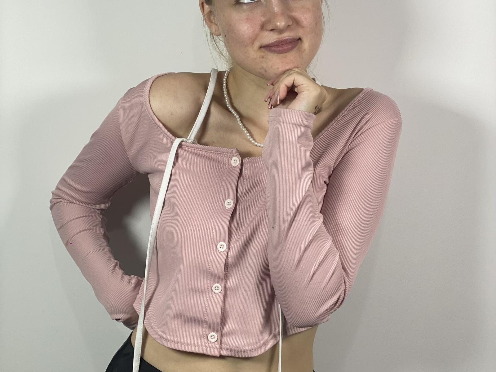 LupitaHeyman con una maglia rosa a maniche lunghe, abbottonata fino in cima, che lascia scoperte le spalle, una spallina bianca, una collana di perle, una mano sul mento, un sorriso appena accennato, pelle chiara, capelli biondi, rossetto rosato..