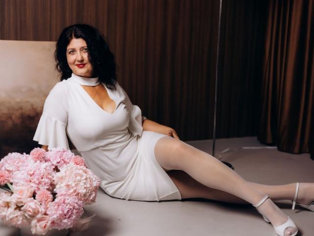 RoseHille with big tits, white dress, choker, and white heels, sitting with legs out, black curly hair, pink flowers in foreground, brown curtain backdrop, smiling.
