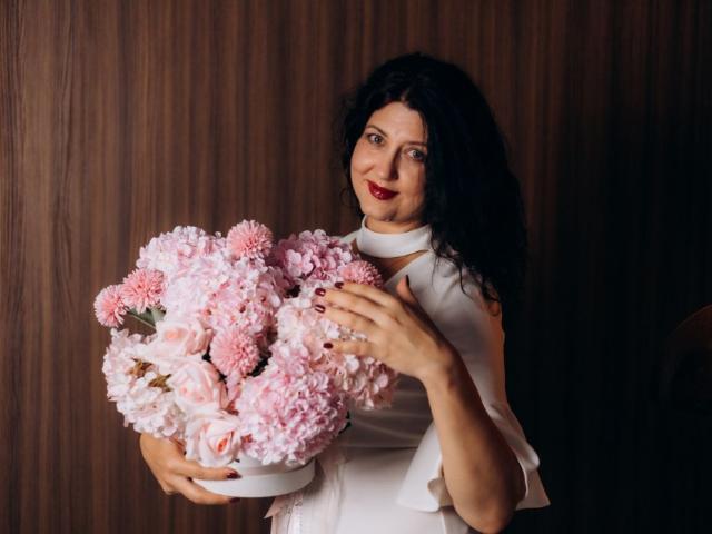 RoseHille with pink and white chrysanthemums, wearing a white sleeveless dress, holding the bouquet with both hands, dark wavy hair, red lipstick, wood-patterned background.