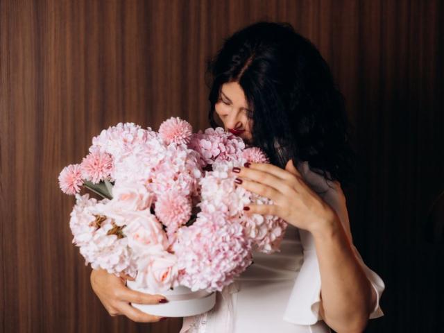RoseHille with curly black hair, holding a bouquet of pink and white flowers, kissing them, wearing a white shirt, nails painted dark red.