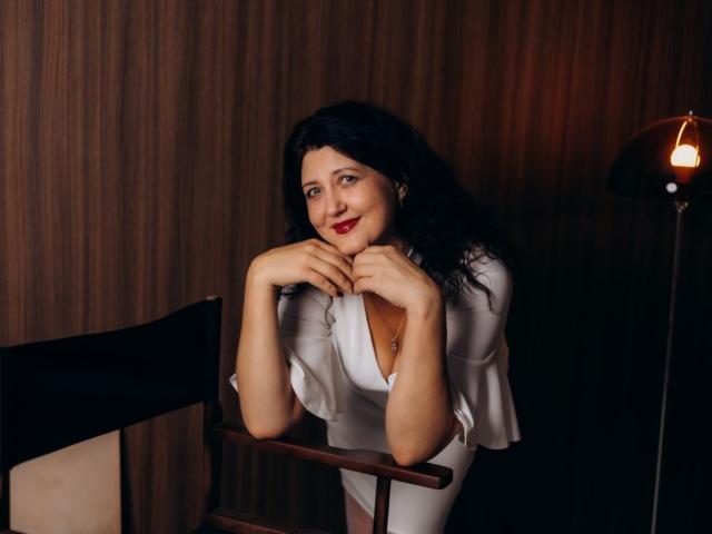 RoseHille leans on a chair, wearing a white dress, hands near her face, smiling with red lipstick, black wavy hair, and a cute nose ring, in a dimly lit room.