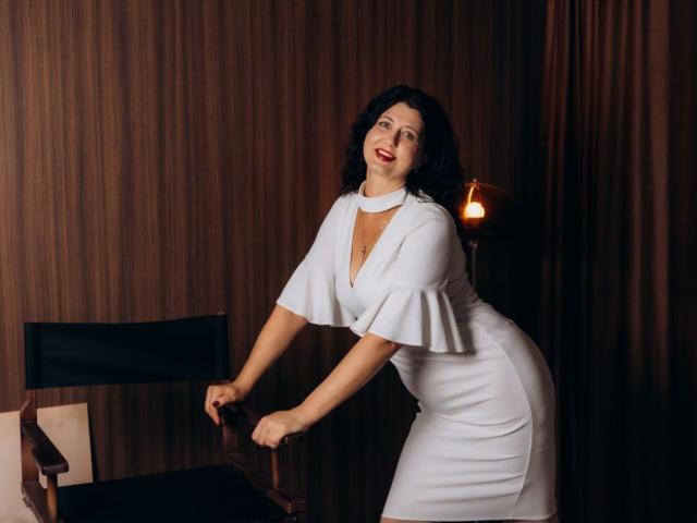 RoseHille, with curly black hair and red lipstick, wearing a white dress with flared sleeves, leans on a director's chair, holding the backrest. Dimly lit, warm glow.