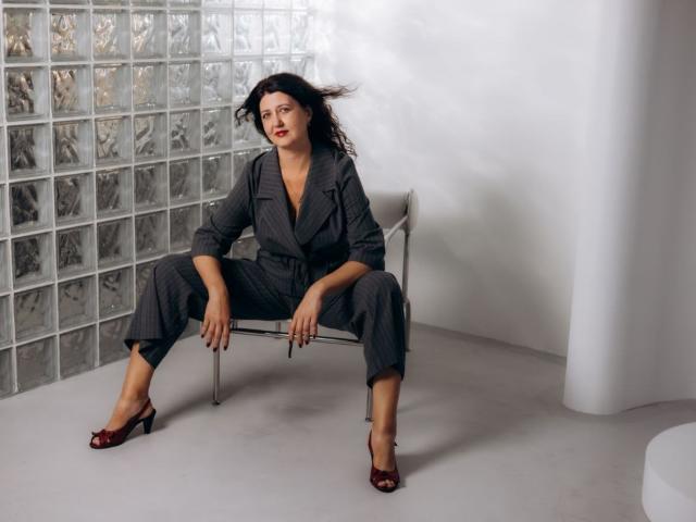 RoseHille with dark hair, wearing a grey pinstripe suit, sitting on a metal chair, legs spread, red open-toe heels, hands on knees, white wall, glass block wall.