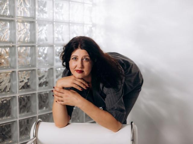 RoseHille leans on a roll of white paper, wearing a gray shirt, with hands crossed, black wavy hair, red lipstick, and a slight smile, in a glass wall setting.
