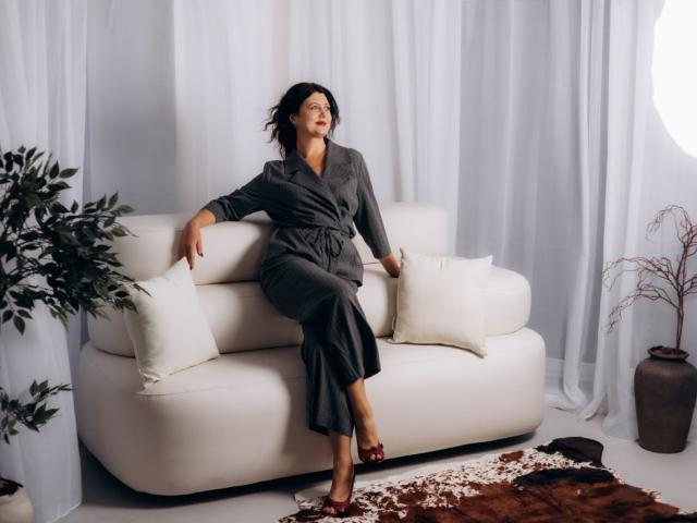 RoseHille with black hair, wearing a gray dress, sitting on a white sofa, red heels, legs crossed, arms resting, white pillows, plant on left, brown rug on floor.
