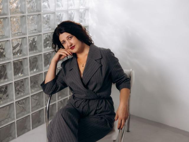 RoseHille with curvy ass, wearing pinstripe gray suit, sitting on chair, black wavy hair, red lipstick, hand on cheek, glass block wall in background, looking at camera.