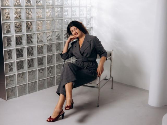 RoseHille sits on a metal chair, wearing a gray pinstripe dress, red high heels, and touching her chin. She has dark curly hair and a confident smile.