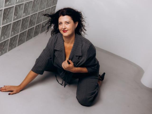 RoseHille with curly black hair, wearing a pinstriped gray blazer and matching pants, kneeling on a gray floor, hand on chest, smiling, red lipstick, black nail polish.