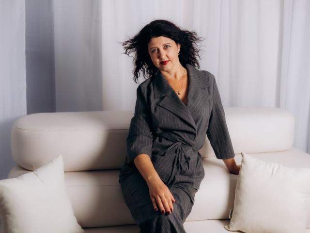 RoseHille with wavy black hair, red lipstick, wearing a gray pinstripe dress, sitting on a white couch with white pillows, looking slightly to the side, hand on her thigh.