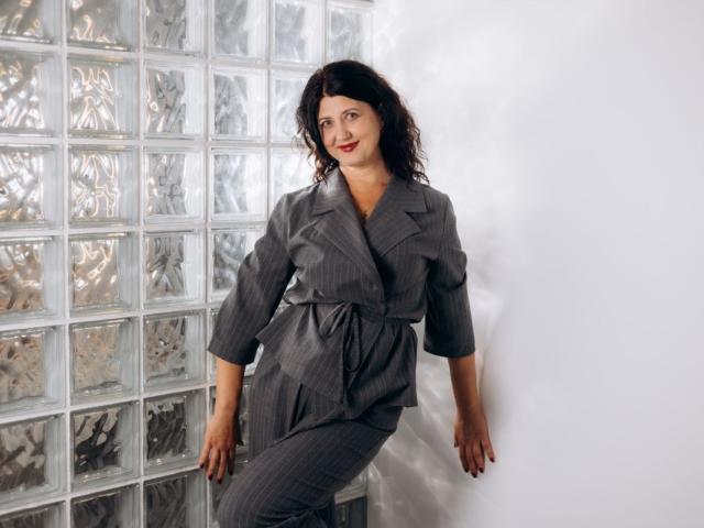RoseHille with wavy dark hair, wearing a gray pinstriped dress, leaning against a white wall, her right hand on the glass block wall, showing a confident smile.