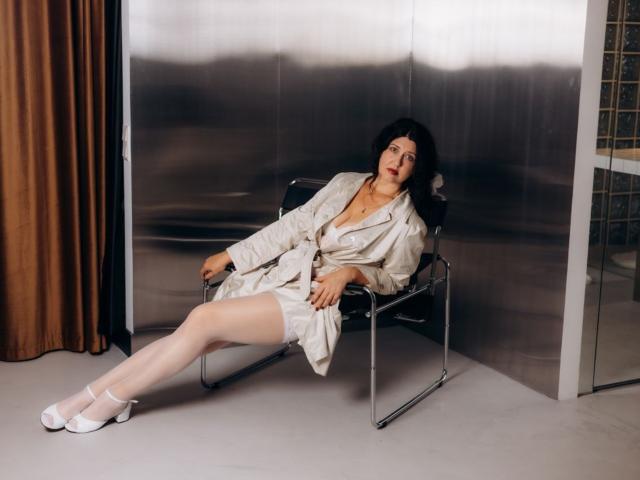 RoseHille is sitting in a chair, wearing a beige trench coat, white stockings, and white heels, with her legs spread, showing her pussy slightly, and a bored expression.