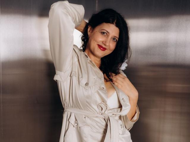 RoseHille with black hair, red lipstick, and white satin blouse, showing tits with her hand on her chest, standing against a blurred metallic background, smiling slightly.