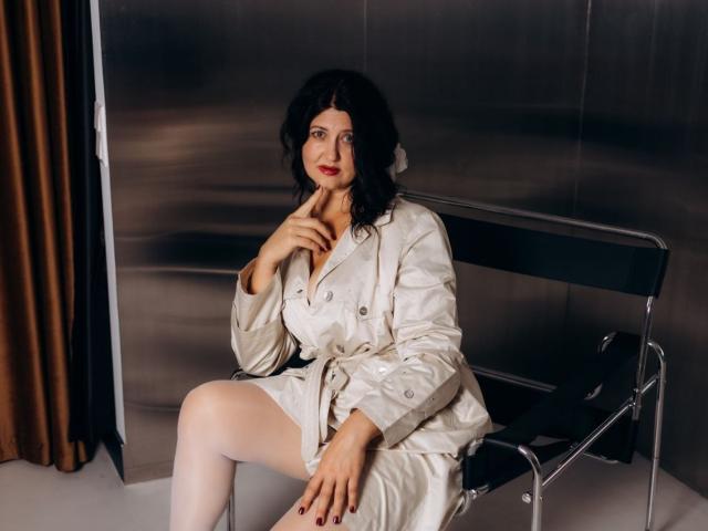 RoseHille sits on a black metal chair, wearing a beige trench coat, white stockings, and red lipstick, with her hand near her chin, looking slightly to the side.