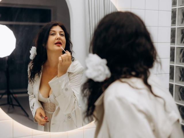 RoseHille with dark curly hair, white button-up shirt, and white flower in hair, applying red lipstick in front of round mirror, holding lipstick in right hand, looking at herself.