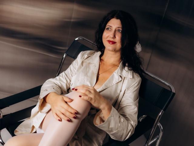 RoseHille with dark curly hair, red lipstick, and white trench coat, sitting on a black chair, hands on her knee, white stockings, red nails, smiling at the camera.