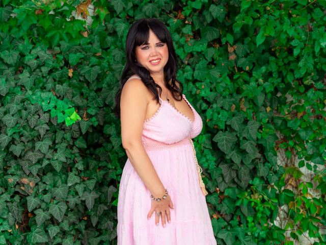 Vivian, uma mulher madura com cabelo preto, vestido rosa e seios visíveis, sorrindo, em pé contra uma parede verde com folhas, com as mãos perto da cintura, usando uma pulseira no pulso direito..