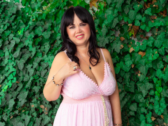 Vivian, uma mulher madura com cabelo preto, vestido rosa e decote, sorrindo, segurando o vestido, fundo com folhagem verde, usando pulseira e colar, em pé, com curvas, tamanho médio de seios, sorrindo..