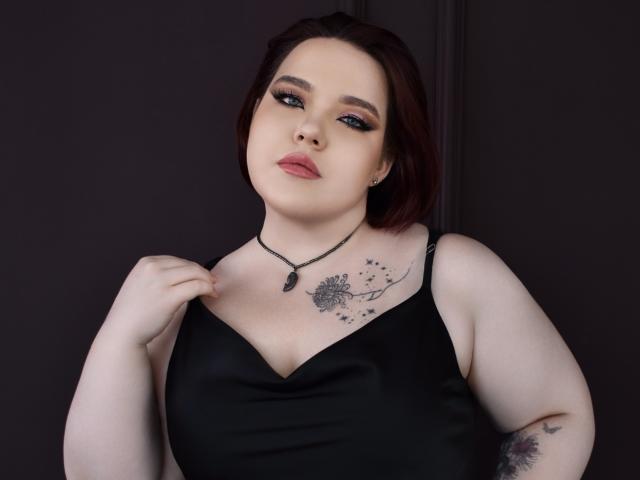 IvoryeFlame has pale skin, red hair, and blue eyes. She's wearing a black dress, holding a strap, showing tattoos on her chest and arm, and has pink lipstick.