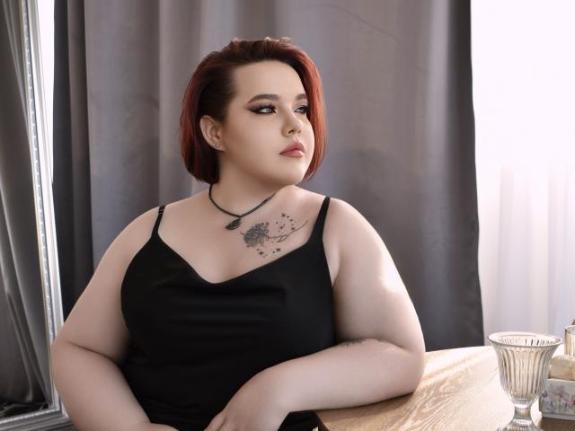 IvoryeFlame with red bob, pale skin, black tank top, showing her tits, tattoo on chest, black necklace, sitting at wooden table, looking to the side, glass on right.
