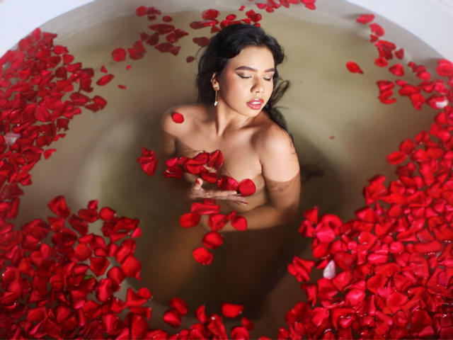NaomiWinters in a bathtub filled with water and red rose petals, holding petals to her tits, with her eyes closed and a calm expression.