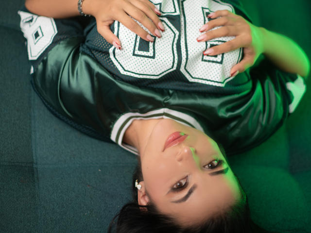 HikariChloe's face is upside down, wearing a green football jersey with number 88, hands on her chest, black hair, green and pink lighting, lying on a dark surface.