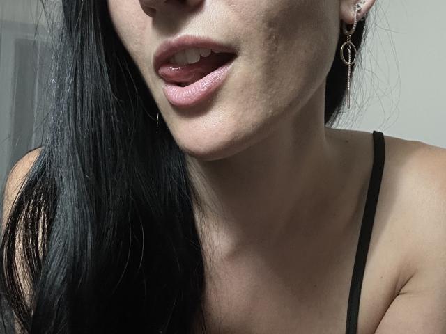 BellaJulls' face, showing her slightly parted pink lips with a hint of her white teeth, black straight hair, and a black spaghetti strap. She's wearing small hoop earrings.
